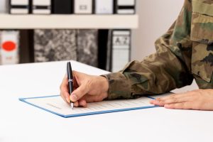 Here is the image of a close-up of a man's hand writing or signing a document on a desk in a military academy.